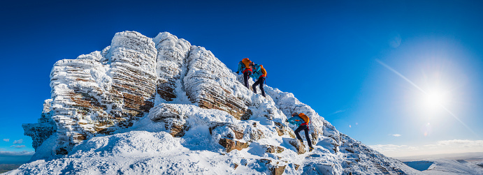 Multiple exposure panorama of teenage female mountaineer climbing snow capped summit peak under deep blue winter skies and bright sunburst. ProPhoto RGB profile for maximum color fidelity and gamut.