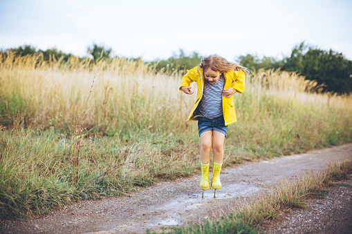 Cute girl wearing yellow rain coat and rubber boots walking in countryside. Jumping on muddy road with puddles. Autumn, yellow grass. Caucasian, blond hair.