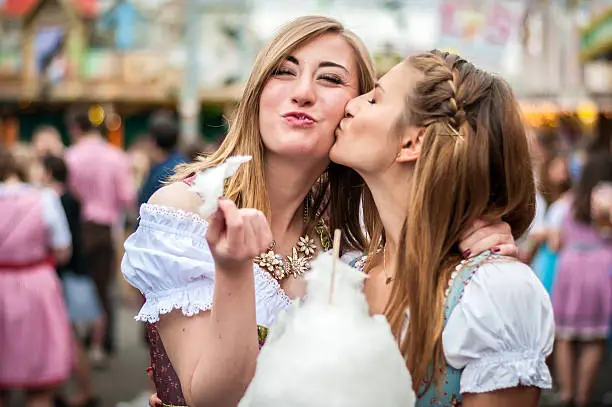 Two young women in traditional Bavarian clothes, dirndl or tracht, kissing with cotton candy floss at the Beer Fest