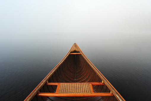 The bow and bow seat of a cedar-strip canoe float on the smooth, still waters of a lake as mist surrounds from every side.