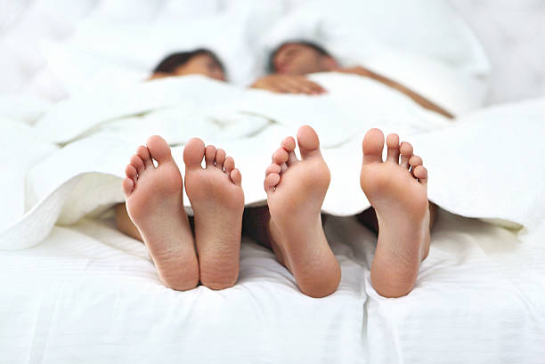 Adult couple talking in bed. Closeup of unrecognizable couple having conversation in bed. They are tucked in, but their feet  stick out of the sheet. Feet are in sharp focus. Front view. protruding stock pictures, royalty-free photos & images