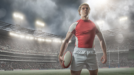 A portrait of a professional muscular male rugby player holding a rugby ball, standing in heroic pose looking into the distance in. The player is wearing a red and white rugby kit and standing on a pitch in a generic floodlit rugby stadium full of spectators under a dramatic stormy sky. 