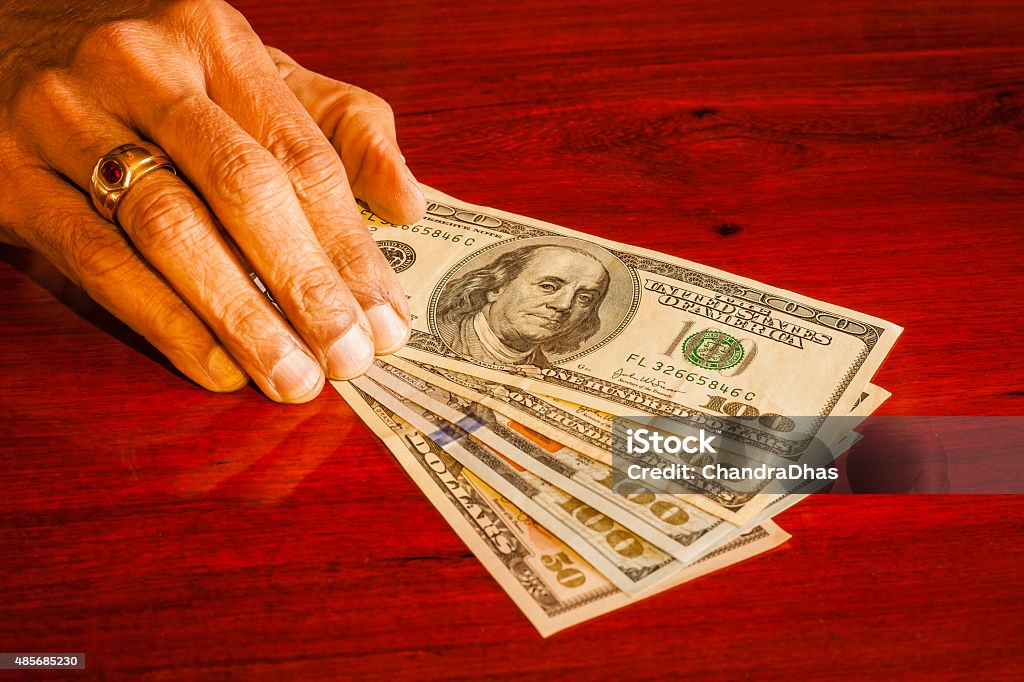 Dollars, USA currency -money on polished rosewood table Senior asian hand puts USA dollars on a polished rosewood table.  Like many asians, he wears a gold ring with a ruby. There are denominations of 100 and a 50 note.  Shot in studio environment; horizontal format. Low-key lighting. Copy space.  Focus on President Franklin's face. 2015 Stock Photo