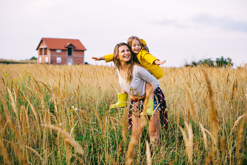 Mother holding, piggybacking her cute daughter, walking through the meadow with house in background. Autumn, yellow grass. Caucasian, blond hair. Wearing rubber boots.