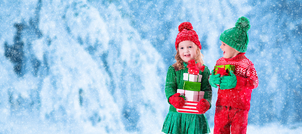 Little girl and boy in red and green knitted hat holding Christmas present boxes in winter park on Xmas eve. Kids play outdoor in snowy winter forest. Children opening presents. Panorama size.