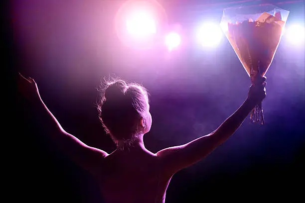 Horizontal composition color photography of young woman artist (singer or classical dancer) receives a bouquet of flowers on stage at the end of the spectacle as a thank you, and saluting the public. Rear view from backstage with spotlight in camera creating lens flare.