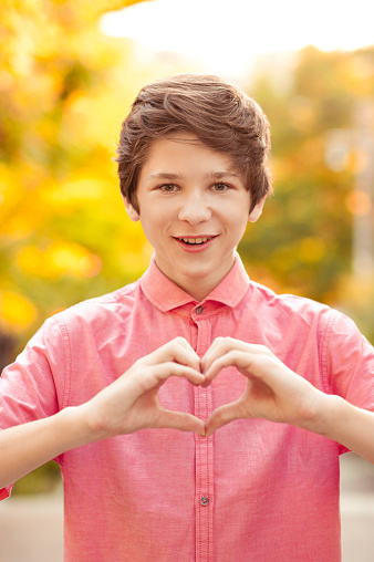 Smiling boy 14-16 year old making heart with fingers outdoors. Looking at camera. Valentines day.