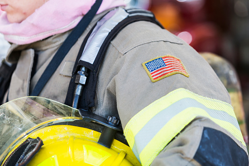 Close up of the arm of a female firefighter in her protective suit.  She is wearing a patch of the American flag on her sleeve, holding a yellow helmet under her arm.  She is cropped so only her chin is visible, so she is unrecognizable.