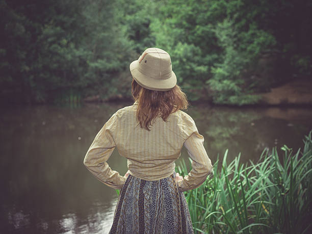 A young woman wearing a safari hat is standing by a pond in the forest on a sunny summer day