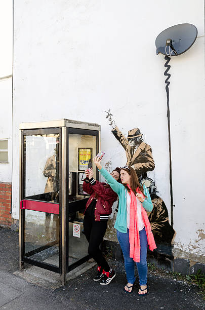 Women pose in front of a possible Banksy artwork, Cheltenham Cheltenham, United kingdom - April 18, 2014: Couple of young women pose for a 'selfie' in front of a possible Banksy artwork. It depicts three government agents monitoring phone conversations from a public phone box.  banksy stock pictures, royalty-free photos & images