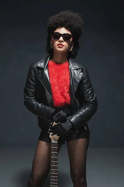 Trendy attractive fashionable young Afro-american lady with a guitar wearing a stylish black leather outfit and sunglasses standing facing the camera, three-quarter pose