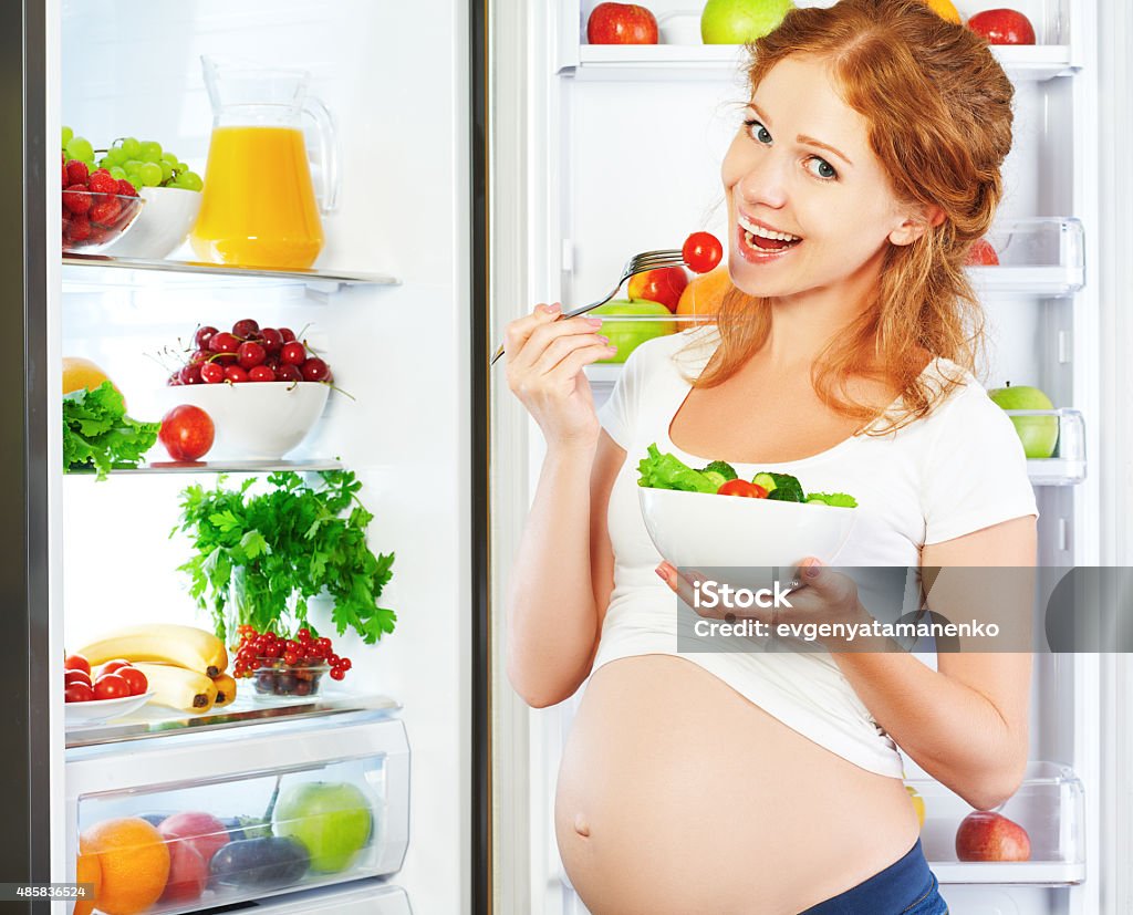 Happy pregnant woman eating salad near refrigerator Happy pregnant woman eating salad near refrigerator at home Kitchen Stock Photo