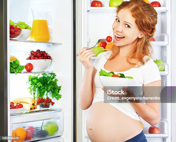 Happy Pregnant Woman Eating Salad Near Refrigerator Stock Photo - Download Image Now