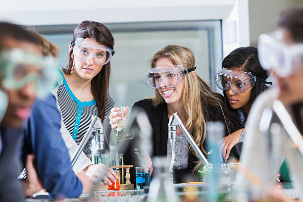 Multi racial group of students in chemistry class Multi-ethnic group of students in chemistry lab, wearing safety goggles, doing science experiment.  Focus on three young women in background. high school high school student science multi ethnic group stock pictures, royalty-free photos & images