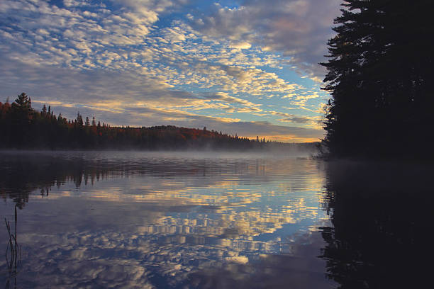 As dawn breaks it colors the sky and an array of magnificent clouds as they reflect on the surface of the calm, misty lake.