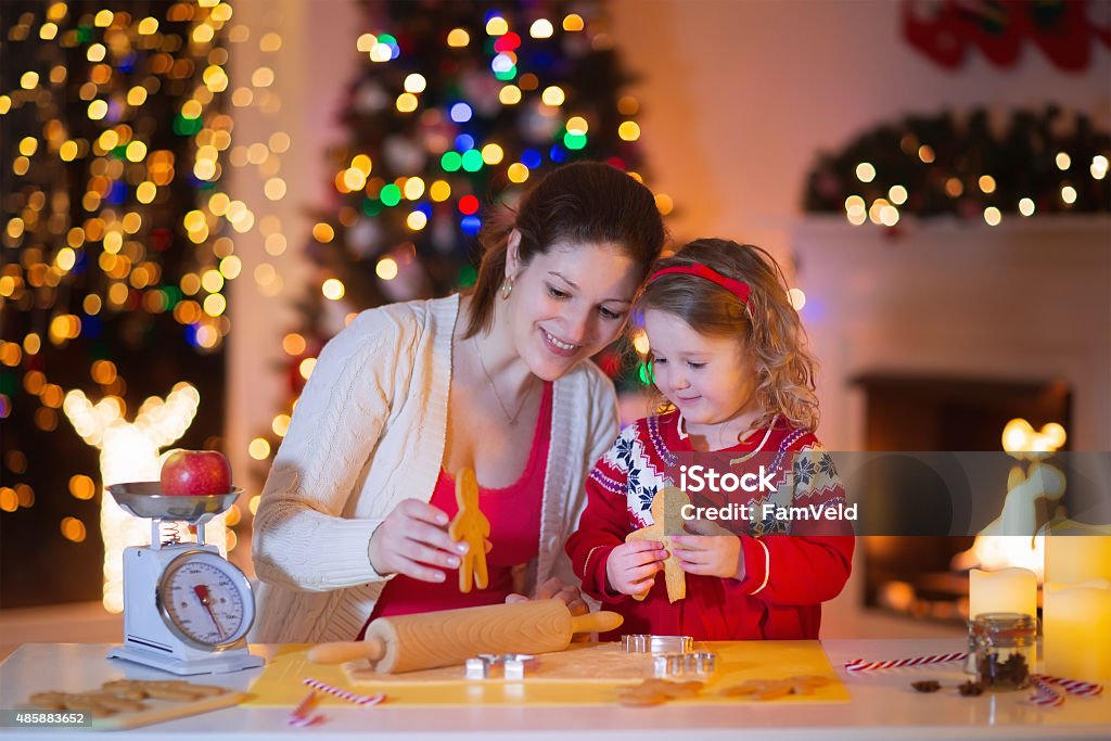 Mother and daughter baking gingerbread for Christmas dinner Mother and little girl baking Christmas pastry. Children bake gingerbread. Toddler child preparing cookie for family dinner on Xmas eve. Decorated kitchen or dining room with fireplace, tree, candles. Adult Stock Photo