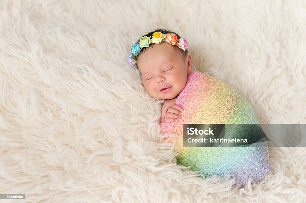 Smiling Newborn Baby Girl Wearing a Rainbow Colored Swaddle A smiling nine day old newborn baby girl bundled up in a rainbow colored swaddle. She is lying on a cream colored flokati (sheepskin) rug and wearing a crown made of roses. Baby - Human Age Stock Photo