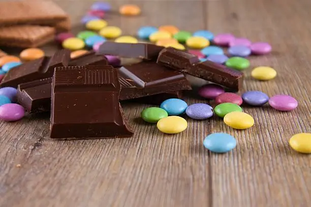 Horizontal photo with heap of dark chocolate pieces on wooden board with colorful chocolate sweet smarties around and biscuits in background.