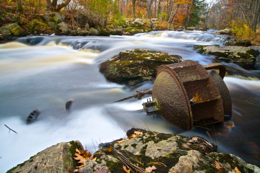 Picture of a rusting water wheel that has fallen into a stream bed.  Some of the original gears are still attached and can be see in the water.