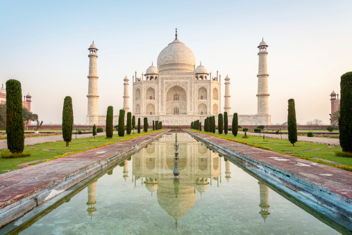 The famous Taj Mahal mausoleum with reflection in the pond, is one of the most recognizable structures worldwide and regarded as one of the eight wonders of the world. Clear blue sky, empty site without people at sunrise. City of Agra, India.