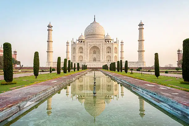 The famous Taj Mahal mausoleum with reflection in the pond, is one of the most recognizable structures worldwide and regarded as one of the eight wonders of the world. Clear blue sky, empty site without people at sunrise. City of Agra, India.