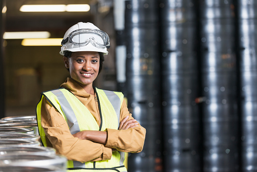 Portrait of a confident, young African American woman working in a manufacturing plant.  She is standing with arms folded, looking at the camera, in a storage facility or warehouse next to stacks of industrial containers.  She is wearing a white hardhat and yellow safety vest.