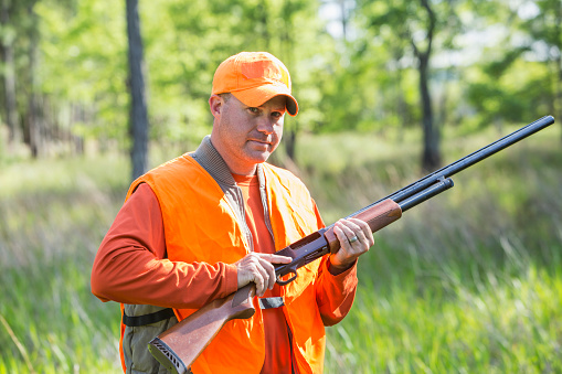A man standing in the woods holding a shotgun looking at the camera.  He is a hunter wearing an orange safety vest and hat, hunting for birds or other small game, or maybe deer.