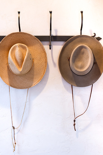 Two Western-style hats (straw and felt) hang on a hat rack against a white plastered wall. Some copy space available.