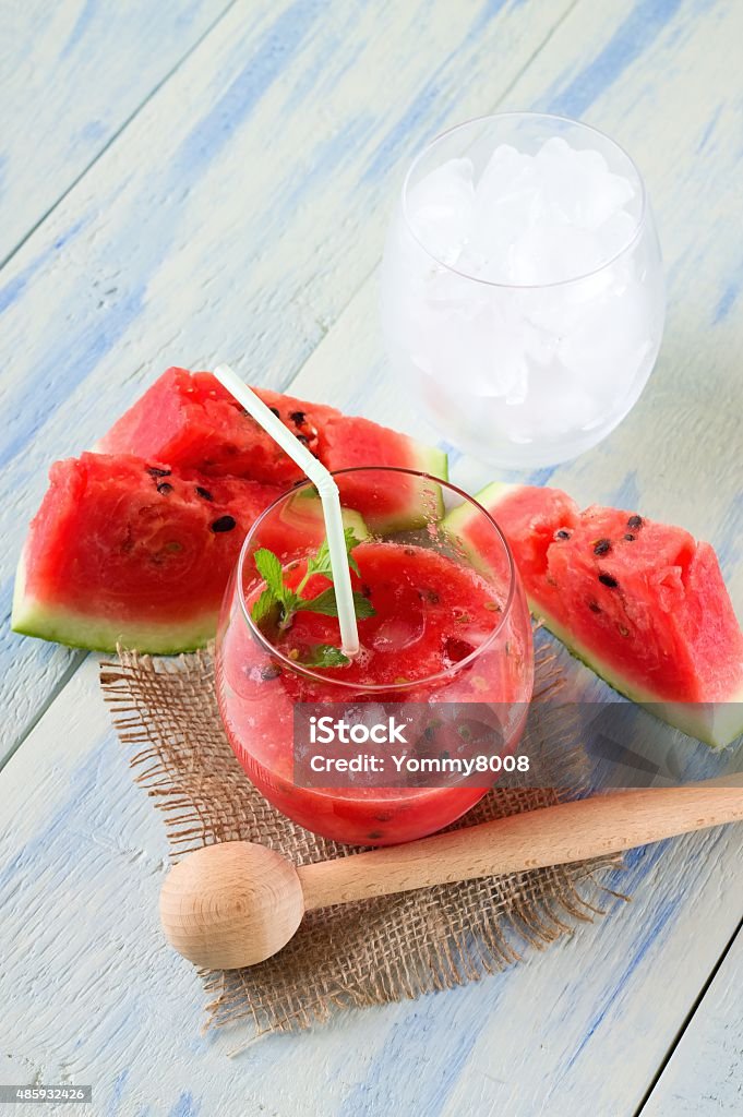 Glass of melon smoothie and glass of ice Vertical photo of two glasses, one with mixed red water melon and second full of ice cubes. Few portions of fruit are around and drink is on piece of burlap. All is on light blue wooden board. 2015 Stock Photo