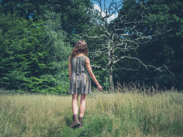 A pretty little girl is picking wildflowers in a lush meadow at sunset. She's alone in a peaceful natural setting, with a forest in the background.