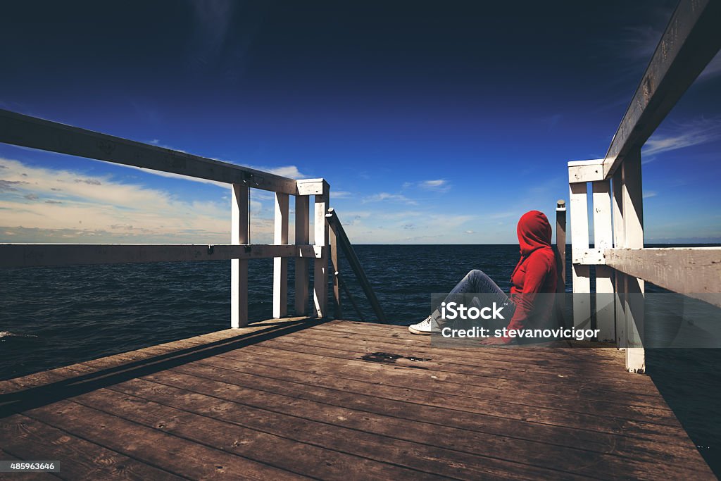 Alone Woman in Red Shirt at the Edge of Pier Alone Young Woman in Red Hooded Shirt Sitting at the Edge of Wooden Pier Looking at Water - Hopelessness, Solitude, Alienation Concept, Retro Toned Hooded Shirt Stock Photo