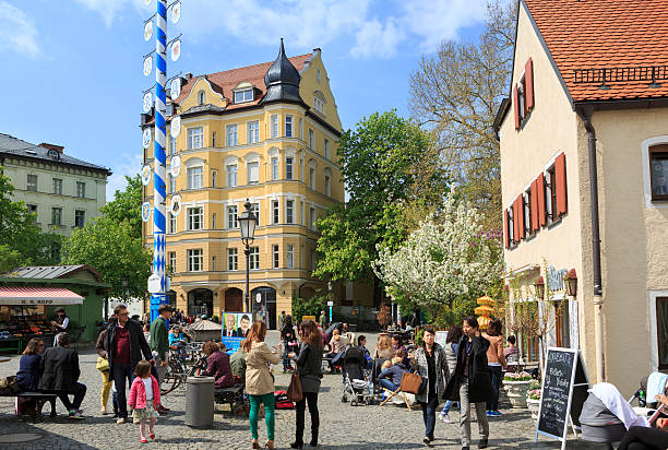 Munich, Germany - April 12, 2014: Several visitors at the square Wienerplatz in the city district Haidhausen in Munich, Bavaria, Germany. On the square there are several cafes and restaurants with outdoor areas and market stalls to the left. A maypole in the centre of the square.