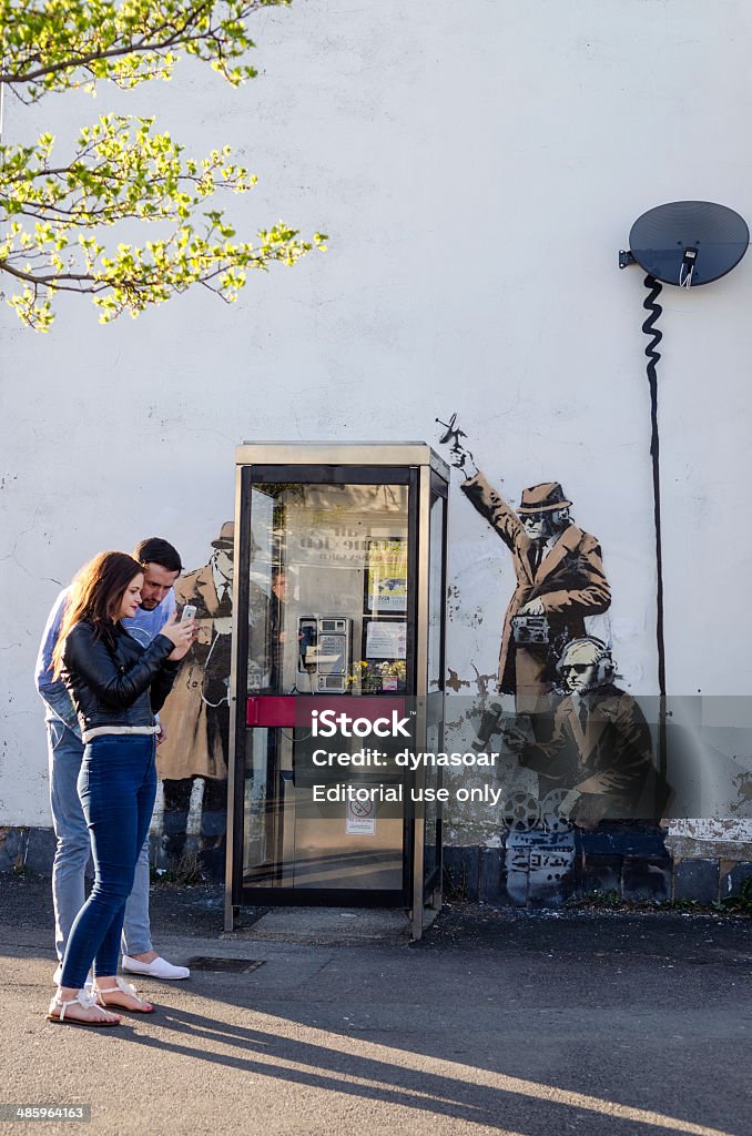 Couple in front of a possible Banksy artwork, Cheltenham Cheltenham, United kingdom - April 18, 2014: Young couple look at photos they've just taken of themselves in front of front of a possible Banksy artwork. The graffiti depicts three government agents monitoring phone conversations from a public phone box.  Banksy Stock Photo