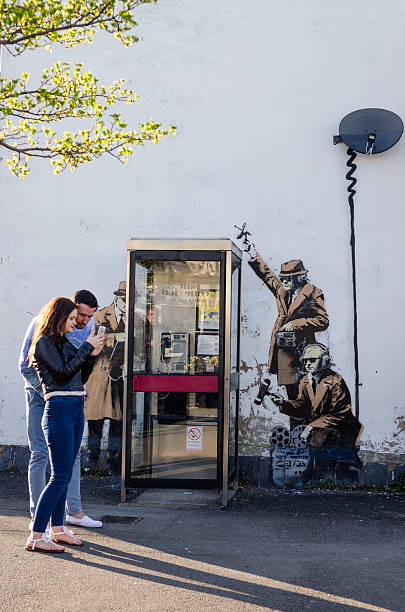 Couple in front of a possible Banksy artwork, Cheltenham Cheltenham, United kingdom - April 18, 2014: Young couple look at photos they've just taken of themselves in front of front of a possible Banksy artwork. The graffiti depicts three government agents monitoring phone conversations from a public phone box.  british telecom photos stock pictures, royalty-free photos & images