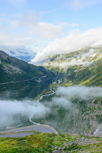 Looking down the Rhone glacier valley, the first kilometres of the Rhone river, towards Gletsch, in the Swiss Alps. Part of the sinuous Furker Pass road can be seen in the foreground; in the distance, clouds are descending the mountainside into the valley floor.