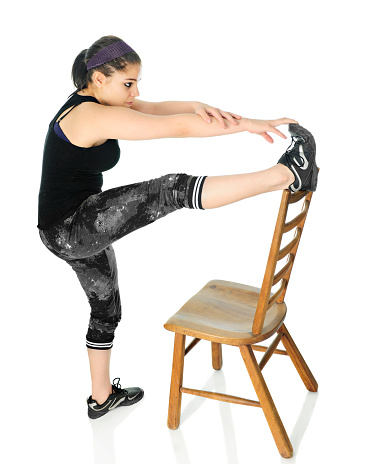 An attractive teen girl working out with one foot on the top of a ladderback chair while stretching to touch her toes.  On a white background.