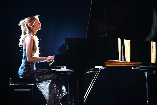 Side view od adult caucasian woman playing piano in a concert. She's placed on a theatre stage and playing grand concert piano. Wearing elegant long dress. Partially backlit, vertical shot. Black stage curtain in background.