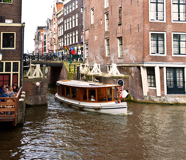 Amsterdam, The Netherlands - July 1, 2012: Canal in Old Centre district of Amsterdam as seen from Oudezijds Voorburgwal. People are taking a sightseeing tour on a boat, eating in outdoor cafe and walking around.