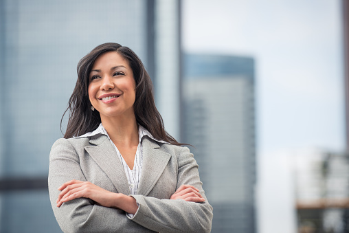 Multi ethnic businesswoman, early thirties, with arms folded standing against downtown buildings.