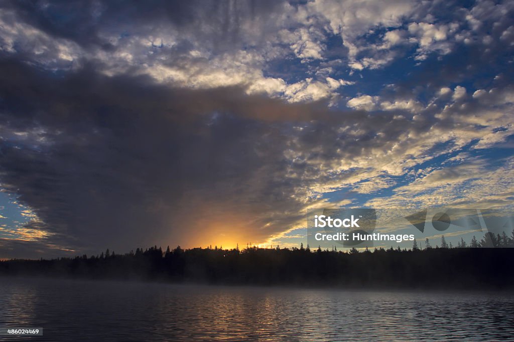 Morning sun shining out from a huge swathe of clouds As dawn breaks it colors the sky and an array of magnificent clouds as they reflect on the surface of the calm, misty lake. Algonquin Provincial Park Stock Photo