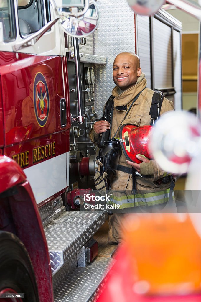 African American fire fighter standing next to truck Portrait of an African American firefighter standing next to a fire truck.  He is wearing a protective suit and holding a red helmet.  He is looking at the camera, smiling. Firefighter Stock Photo