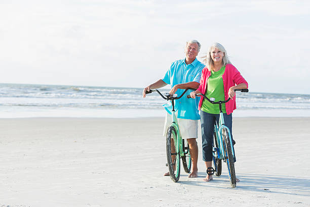 matura coppia biciclette equitazione sulla spiaggia - mature adult bicycle senior adult heterosexual couple foto e immagini stock