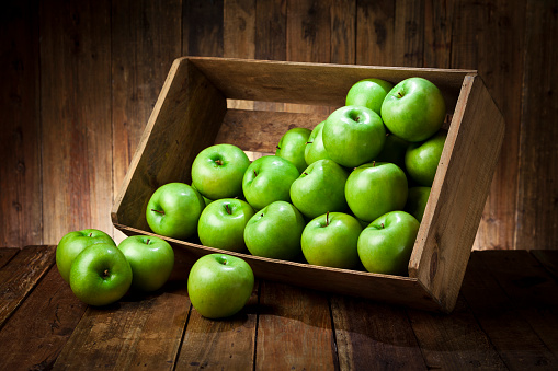 Side view of a tilted wooden crate filled with fresh organic green apples sitting on rustic wood table. The crate is tilted with some apples out of it.Predominant color is brown and green. Low key DSRL studio photo taken with Canon EOS 5D Mk II and Canon EF 24-105mm f/4L IS USM Lens