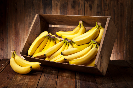 Side view of a tilted wooden crate filled with fresh organic bananas sitting on rustic wood table. The crate is tilted with some bananas out of it. Predominant color is brown and yellow.Low key DSRL studio photo taken with Canon EOS 5D Mk II and Canon EF 24-105mm f/4L IS USM Lens