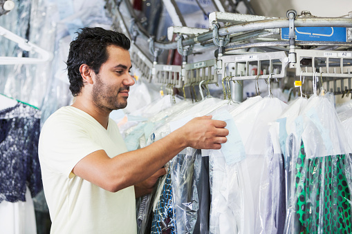 An Hispanic man working in a dry cleaning store, reaching into a rack of drycleaned clothing hanging on hangers and covered in plastic.  He is wearing a white t-shirt.
