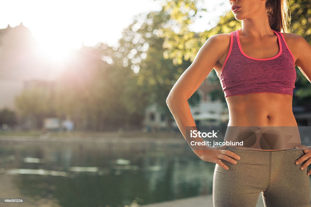 Young woman in sportswear with fit body Cropped shot of fit young woman in sportswear standing with her hands on hips outdoors. Female runner ready for a run with sun flare. Women Stock Photo