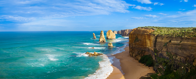 Panoramic image of morning light on the landmark Twelve Apostles along the Great Ocean Road in Victoria, Australia. There is some cloud in the blue sky and the sea reflects a turquoise colour.