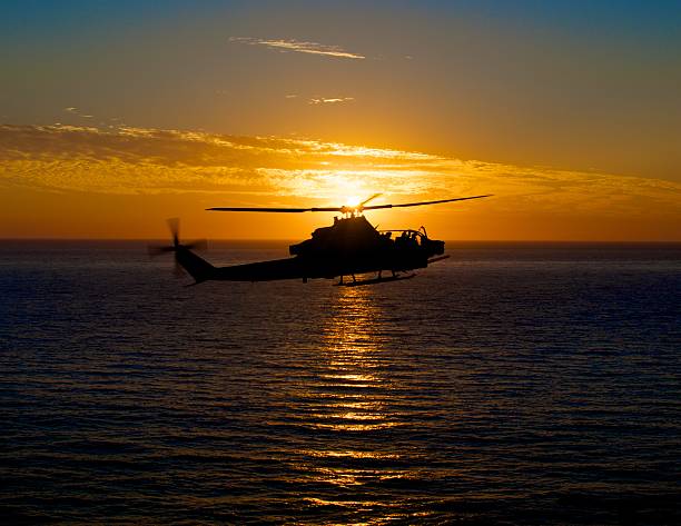 silhouette of AH-1 Super Cobra Helicopter flying over ocean in front of a sunset