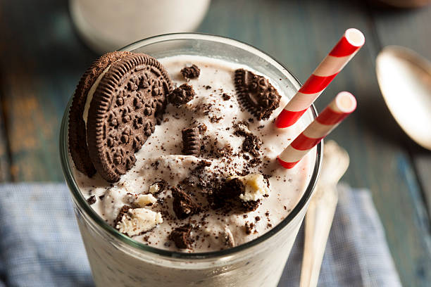 Milkshake with whole cookie and crumbs A cookies and cream milkshake sits on a wooden table in a tall glass with a spoon beside it.  There is a large chocolate sandwich cookie in the milkshake along with crumbled chocolate cookies.  There are two old-fashioned red-and-white straws standing in the glass. chocolate shake stock pictures, royalty-free photos & images