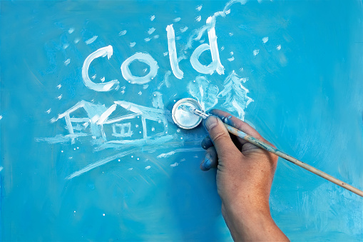 Artist hand with paintbrush writing word Cold and painting winter landscape in childish manner on the blue board, concept winter